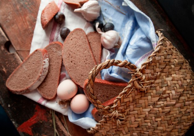 Cesta de vista superior com fatias de pão ovos, ameixa e alhos ao redor em uma mesa de madeira.