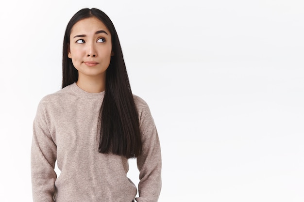 Cética, insegura mulher asiática atraente com longos cabelos escuros, tem dúvidas e hesitações, fazendo escolha com expressão incerta, sorriso afetado olhar suspeito canto superior direito, preocupado