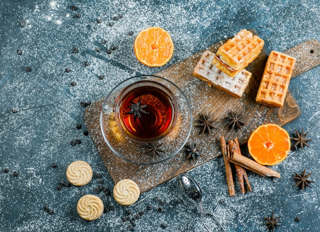 Chá com waffle, biscoito, especiarias, chips de chocolate, filtro, laranja em um copo na superfície azul e placa de corte, plana leigos.