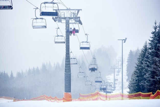 Chairlift usando para transportar passageiros para cima e para baixo nas montanhas durante a queda de neve. densa floresta de abetos coberta de neve ao redor.