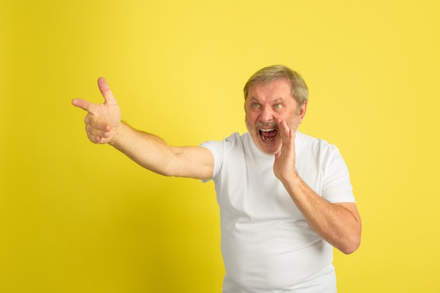 Chamando, apontando. Retrato de homem caucasiano isolado em fundo amarelo do estúdio. Lindo modelo masculino posando de camisa branca. Conceito de emoções humanas, expressão facial, vendas, anúncio. Copyspace.