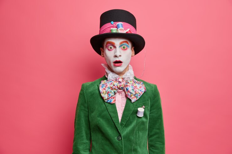 Foto grátis chapeleiro chocado e envergonhado olha para a câmera com olhos arregalados, vestido com uma jaqueta verde gravata borboleta e um chapéu grande com maquiagem colorida participa de performance isolada sobre uma parede rosada