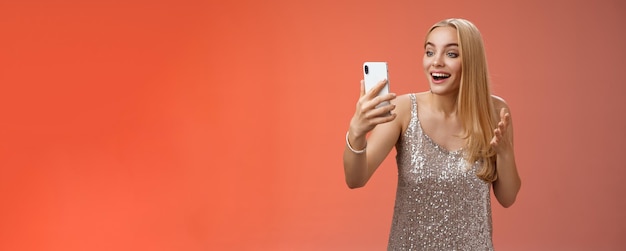 Foto grátis charmosa elegante bonita garota loira em vestido prateado falando chamada de vídeo falando olhando smartphone