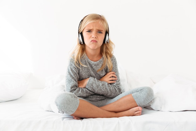 Chateado criança feminina em fones de ouvido, sentado com os braços cruzados e pernas na cama