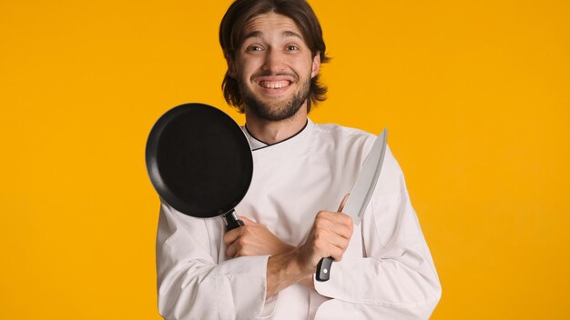 Chef atraente vestindo uniforme segurando faca e frigideira, mantendo as mãos cruzadas sobre fundo colorido Jovem sorridente pronto para cozinhar