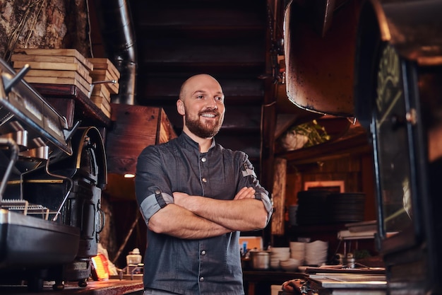 Chef confiante vestindo uniforme posando com os braços cruzados e desviar o olhar em uma cozinha de restaurante.