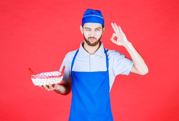 Chef de avental azul segurando uma cesta de pão coberta com uma toalha vermelha e mostrando sinal de prazer.