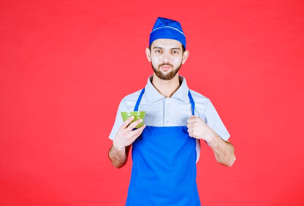 Chef de avental azul segurando uma xícara de cerâmica verde e mostrando sinal de satisfação.