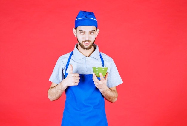 Chef de avental azul segurando uma xícara de cerâmica verde e mostrando sinal de satisfação.