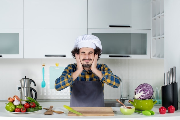 Chef do sexo masculino nervoso com legumes frescos e cozinhando com utensílios de cozinha e fechando os ouvidos na cozinha branca