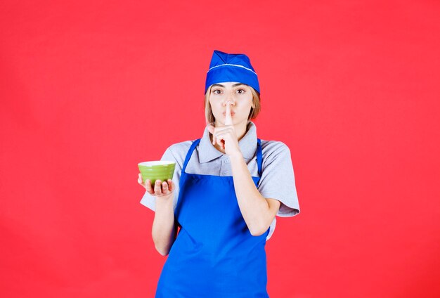 Chef feminina de avental azul segurando uma xícara de macarrão verde e pedindo silêncio