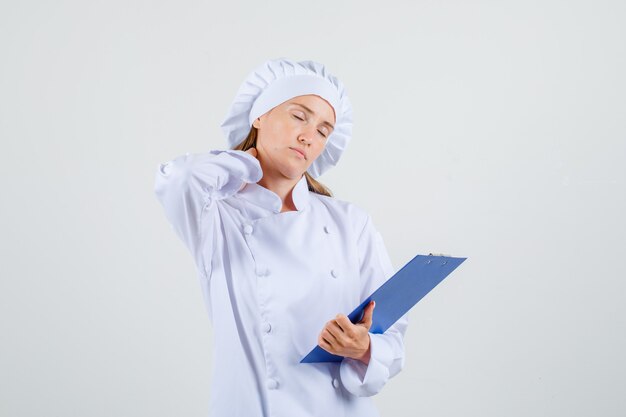 Chef feminina de uniforme branco segurando uma prancheta com a mão no pescoço e parecendo cansada