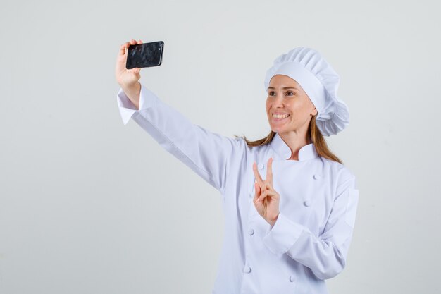 Chef feminina de uniforme branco tirando selfie com o sinal V e parecendo alegre