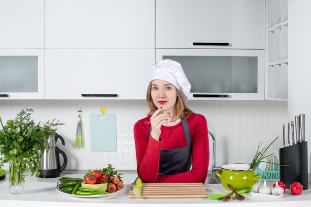 Chef feminina de uniforme em pé atrás da mesa da cozinha com a mão no queixo