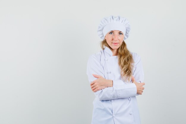 Chef feminina em pé com os braços cruzados, em uniforme branco e parecendo confiante