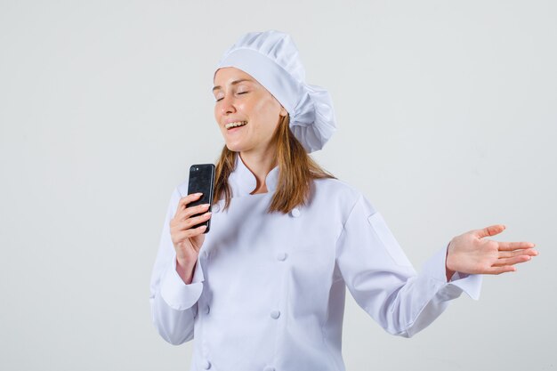 Chef feminina segurando um smartphone com a mão aberta em uniforme branco e parecendo alegre
