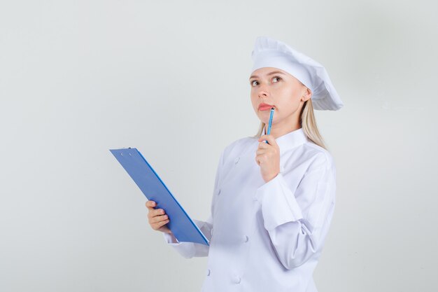 Chef feminina segurando uma prancheta com um lápis no uniforme branco e parecendo pensativa