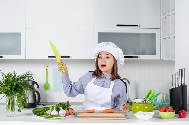 Chef feminina surpresa e legumes frescos com equipamento de cozinha e apontando algo com uma faca na cozinha branca