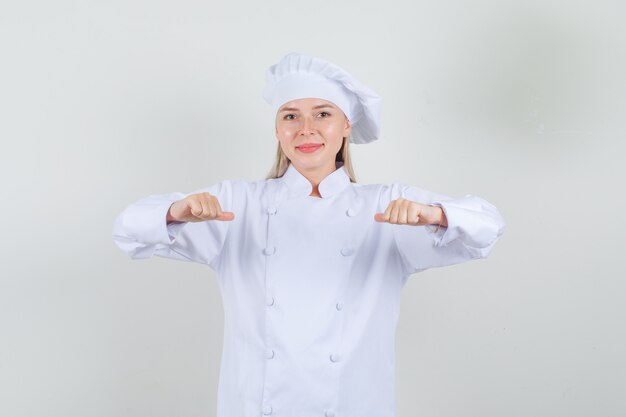 Chef feminino apontando os polegares para si mesma em uniforme branco e parecendo alegre.