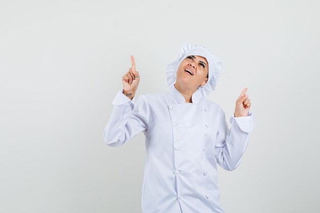 Chef feminino apontando para cima em uniforme branco e parecendo um sonho.