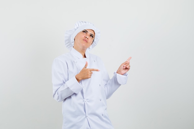 Chef feminino apontando para o lado e para cima em uniforme branco e parecendo hesitante.