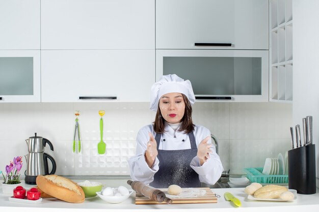 Chef feminino de uniforme em pé atrás da mesa, manchando o rosto com farinha na cozinha branca