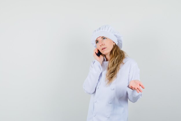 Chef feminino falando no celular em uniforme branco e parecendo confuso.