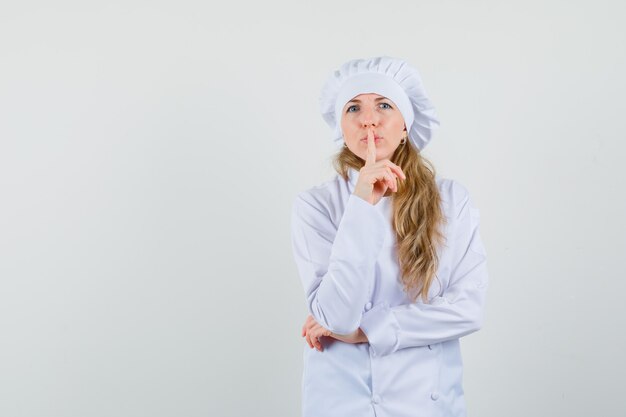 Chef feminino mostrando gesto de silêncio em uniforme branco e olhando com cuidado.