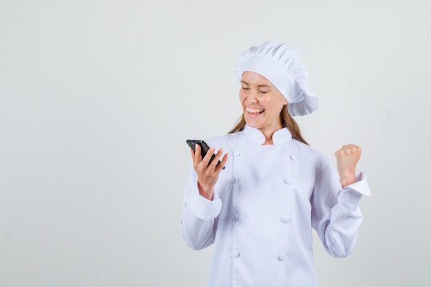 Chef feminino mostrando o gesto do vencedor enquanto segura o smartphone em uniforme branco. vista frontal.