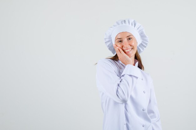 Chef feminino segurando a mão na bochecha em uniforme branco e parecendo feliz. vista frontal.