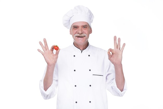 Chef idoso profissional com uniforme branco e chapéu, segurando tomate enquanto permanece na parede branca