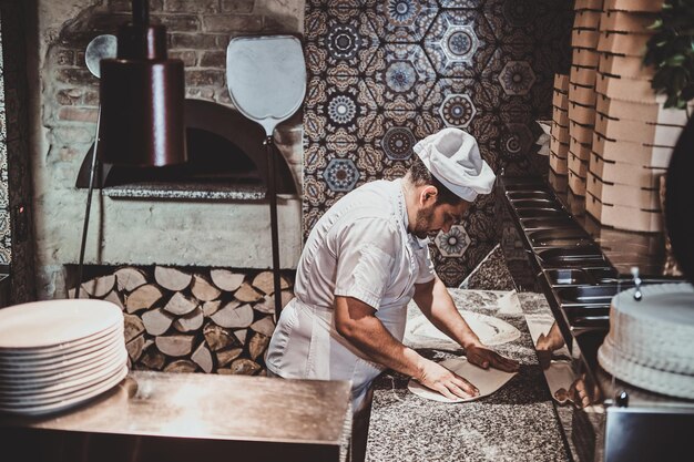Chef italiano de uniforme está preparando pastelaria para pizza na cozinha.