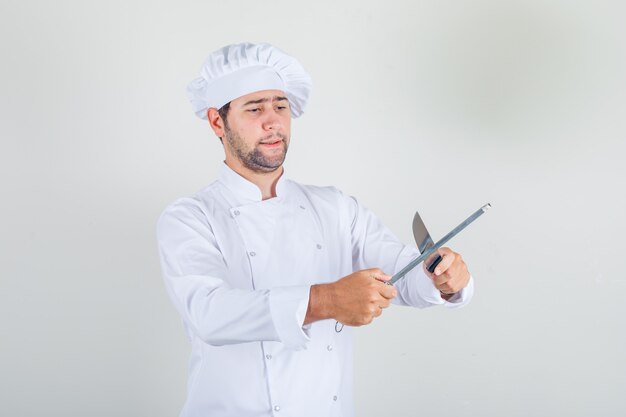 Chef masculino afiando faca em uniforme branco e parecendo ocupado