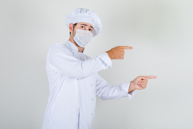 Chef masculino com uniforme branco, máscara médica apontando os dedos e olhando com cuidado