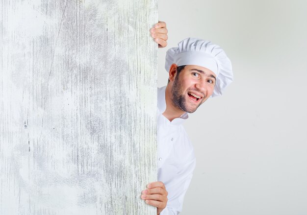 Chef masculino com uniforme branco olhando por trás de uma placa de madeira e parecendo alegre