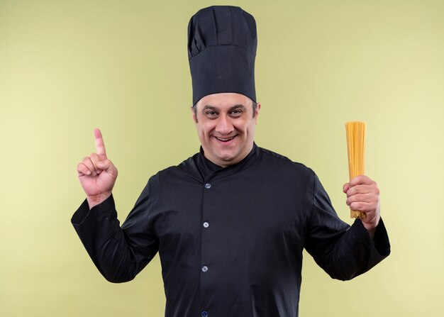 Chef masculino cozinheiro vestindo uniforme preto e chapéu de cozinheiro segurando espaguete apontando com o dedo para cima, olhando para a câmera, sorrindo com carinho em pé sobre fundo verde