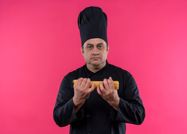 Chef masculino cozinheiro vestindo uniforme preto e chapéu de cozinheiro segurando espaguete cru olhando para a câmera wirth rosto sério em pé sobre fundo rosa