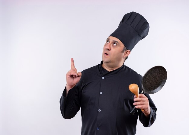 Chef masculino cozinheiro vestindo uniforme preto e chapéu de cozinheiro segurando uma panela e uma colher de pau apontando com o dedo indicador para cima surpreso em pé sobre um fundo branco