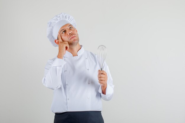 Chef masculino de uniforme, avental e chapéu segurando o batedor, pensando e olhando com cuidado