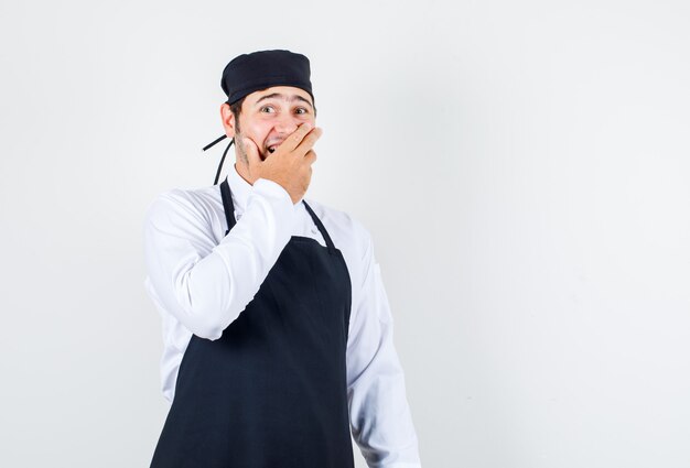 Chef masculino de uniforme, avental segurando a mão na boca e olhando surpreso, vista frontal.