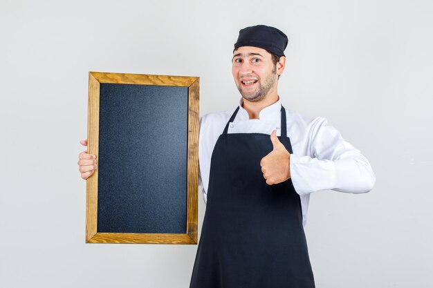 Chef masculino de uniforme, avental segurando uma lousa com o polegar para cima e olhando feliz, vista frontal.