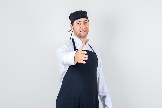 Chef masculino estendendo a mão para saudação de uniforme, avental e parecendo jovial. vista frontal.