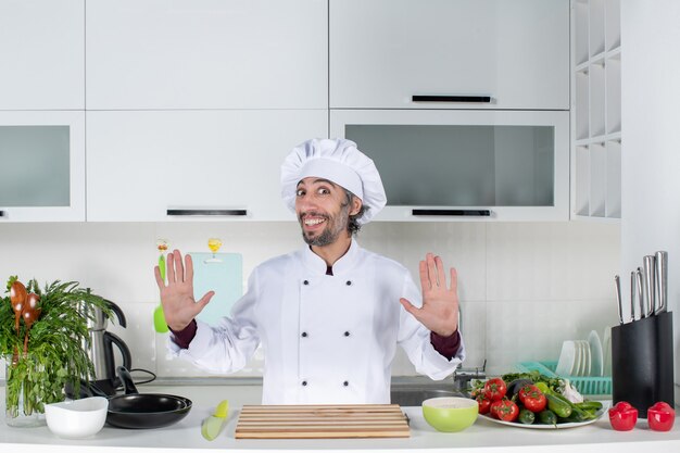 Chef masculino feliz de vista frontal de uniforme em pé atrás da mesa da cozinha na cozinha moderna