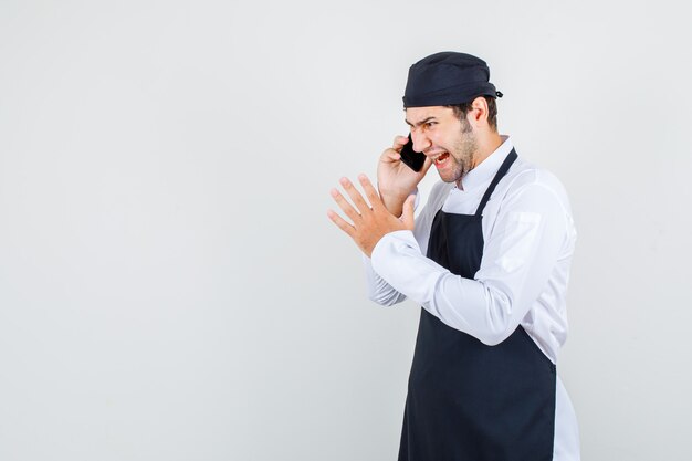 Chef masculino gritando enquanto fala no smartphone de uniforme, avental e olhando furioso, vista frontal.