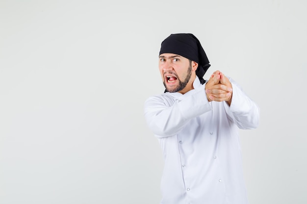 Chef masculino mostrando gesto de arma apontado em uniforme branco e olhando com raiva, vista frontal.