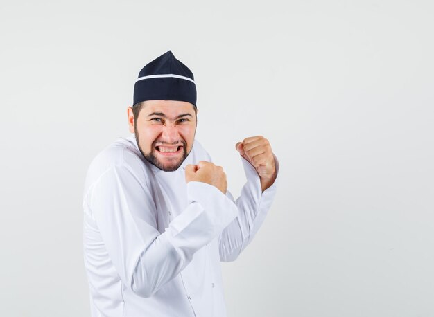 Chef masculino mostrando gesto de vencedor em uniforme branco e olhando feliz, vista frontal.