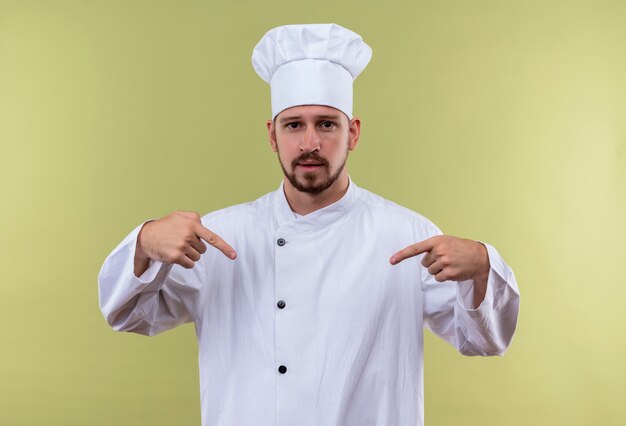 Chef masculino profissional satisfeito com uniforme branco e chapéu de cozinheiro apontando para si mesmo com as duas mãos em pé sobre o fundo verde