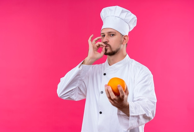 Chef masculino profissional satisfeito cozinheiro de uniforme branco e chapéu de cozinheiro segurando uma fruta laranja fresca mostrando sinal de estar em pé delicioso sobre fundo rosa