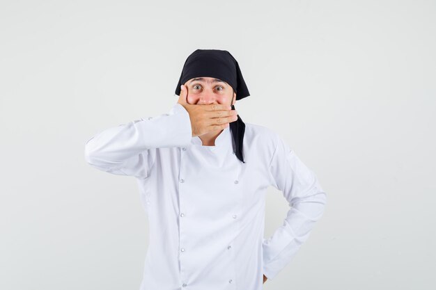 Chef masculino segurando a mão na boca em uniforme branco e olhando surpreso, vista frontal.