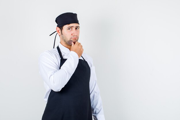 Chef masculino segurando a mão no queixo de uniforme, avental e olhando pensativo. vista frontal.